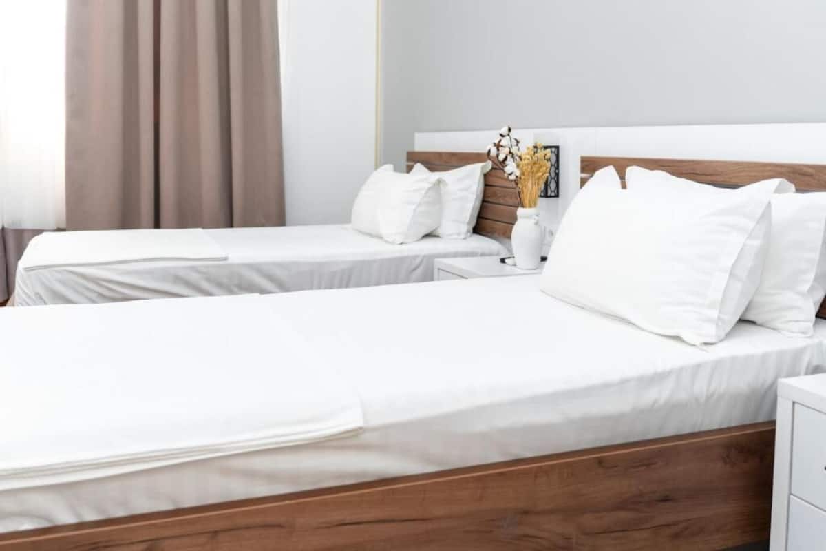 A hotel room with two single beds, a wooden headboard, a bedside table with a vase of flowers, and a window with curtains.