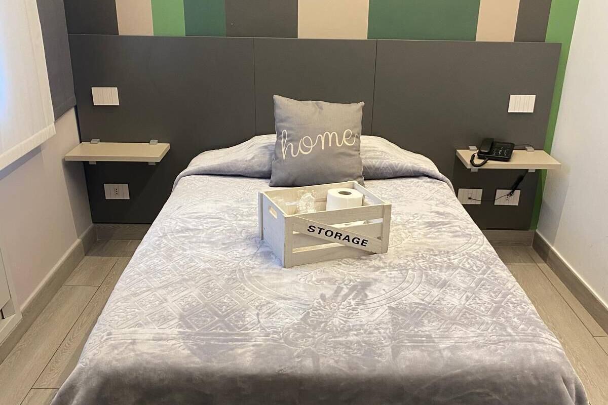 Un lit bien fait, avec une couette grise, un oreiller orné du mot « home » et une boîte de rangement en bois étiquetée « RANGEMENT », le tout posé sur un plancher en bois.