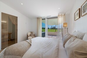 Ein Schlafzimmer mit einem großen Bett, einem Sessel, einem Tisch und Blick durch eine Schiebetür auf einen Außenbereich.