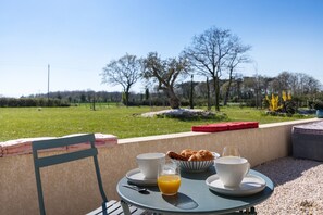 Ein Tisch mit Frühstücksangeboten, ein Stuhl und Blick auf ein Wiesfeld und Bäume.