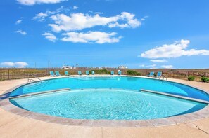 A large, curved swimming pool with clear blue water, surrounded by a low stone wall and a metal fence. - Beachside Townhome | Pool, Gulf Views & Beach Path (Corpus Christi)