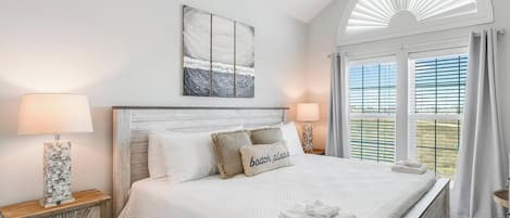 A bedroom with a large bed, two bedside tables, a ceiling fan, a window with shutters, and a beach-themed pillow.