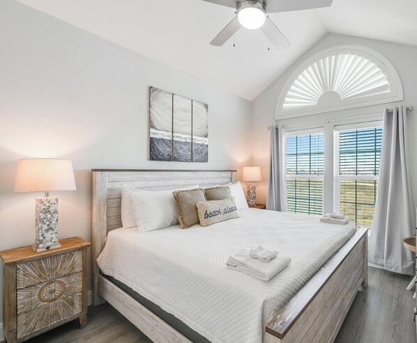 A bedroom with a large bed, two bedside tables, a ceiling fan, a window with shutters, and a beach-themed pillow. - Beachside Townhome | Pool, Gulf Views & Beach Path (Corpus Christi)