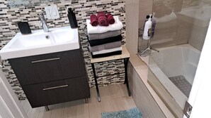 Un baño moderno con lavamanos, mueble de madera oscura, una mesita con toallas y zona de ducha.