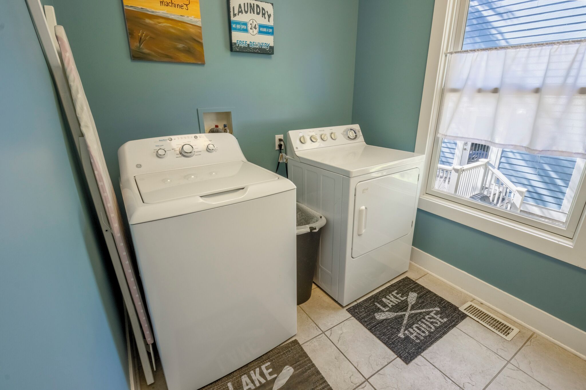 A laundry room with two white washing machines, a small trash bin, and a window with a view of a balcony.