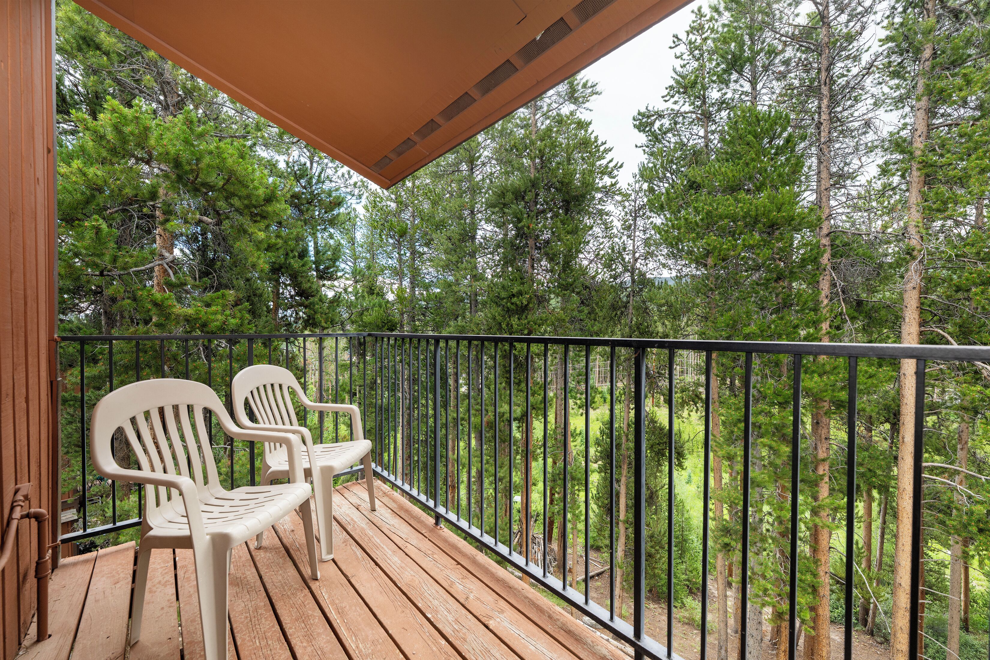 Um deck de madeira com duas cadeiras brancas e um guarda-corpo preto, com vista para uma floresta.