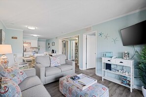 A living room with a grey sofa, a matching ottoman, a white console table with decorative items, and a flat-screen TV mounted on the wall. - Resort View - Tennis Villas E14-Professionally Managed by HHIB&T Vacation Rentals (Hilton Head Island)