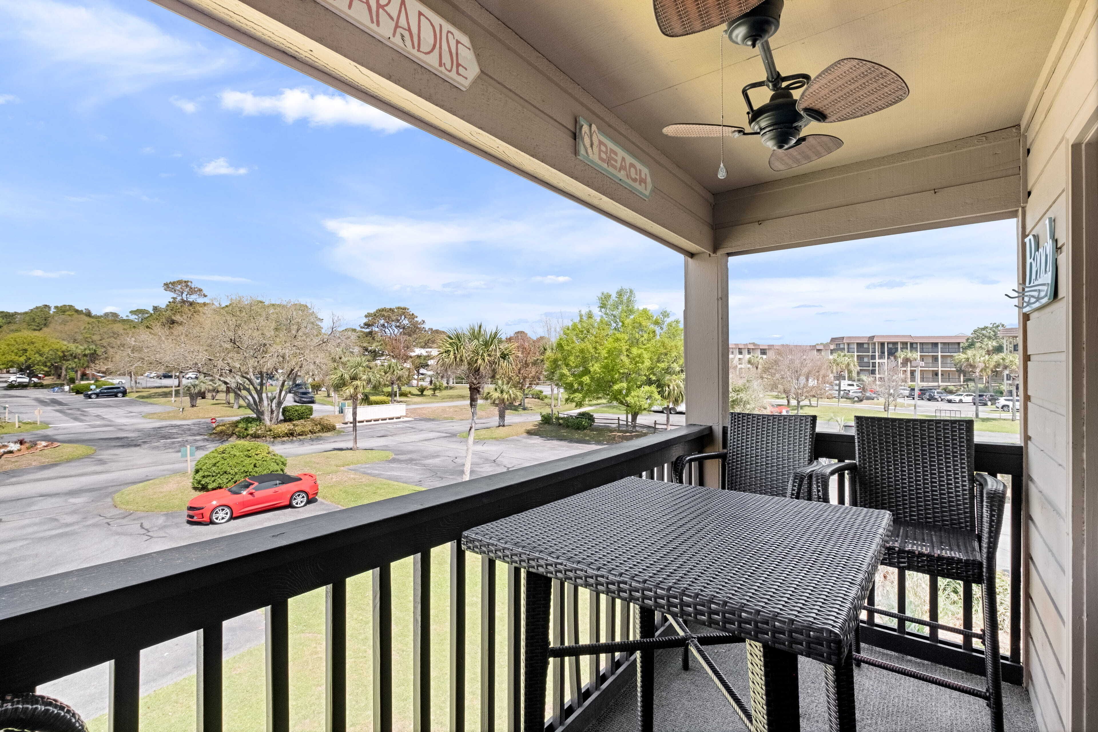 A balcony with wicker furniture, a ceiling fan, and a view of a parking lot and buildings.