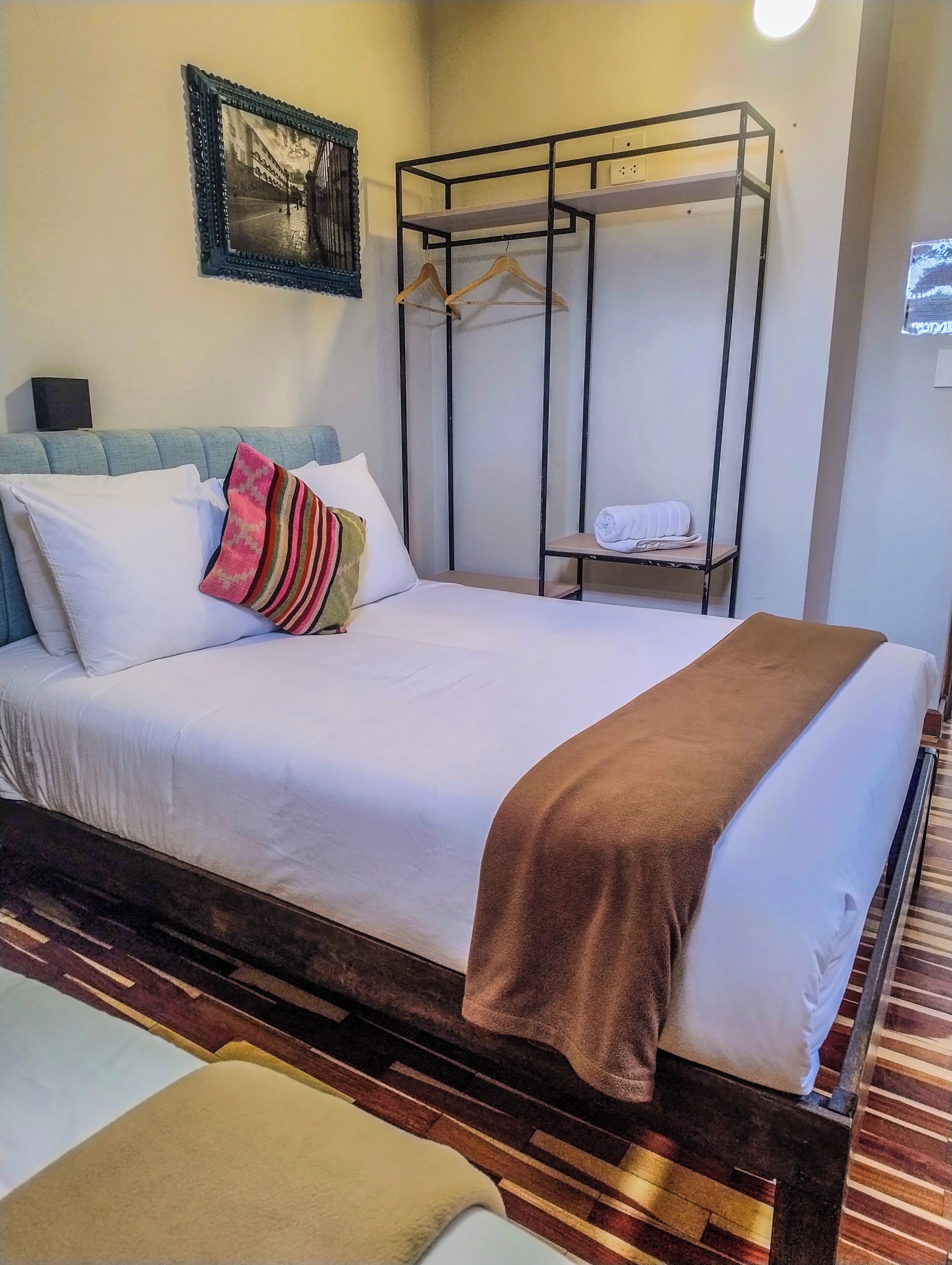 A hotel room with a bed, a hanging clothes rack, a framed picture on the wall, and a small table with a towel.