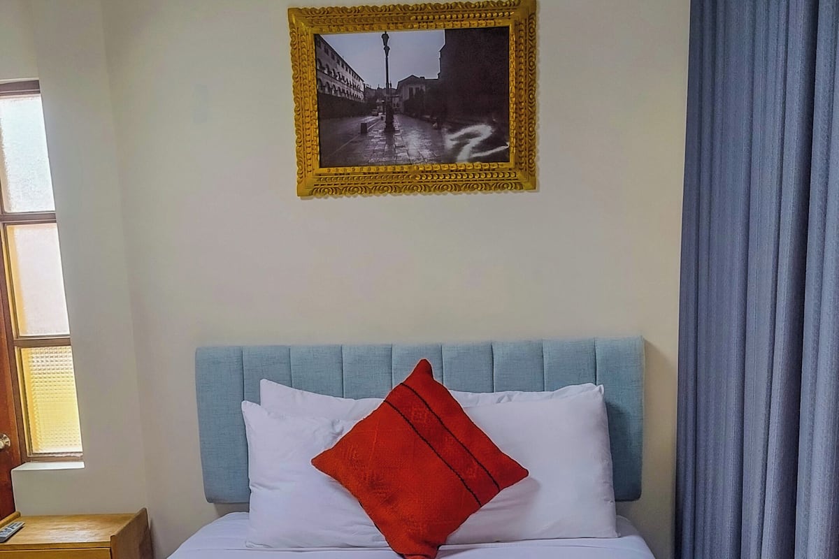 A bed with a headboard, a pillow, a nightstand, a window, and a framed picture on the wall.