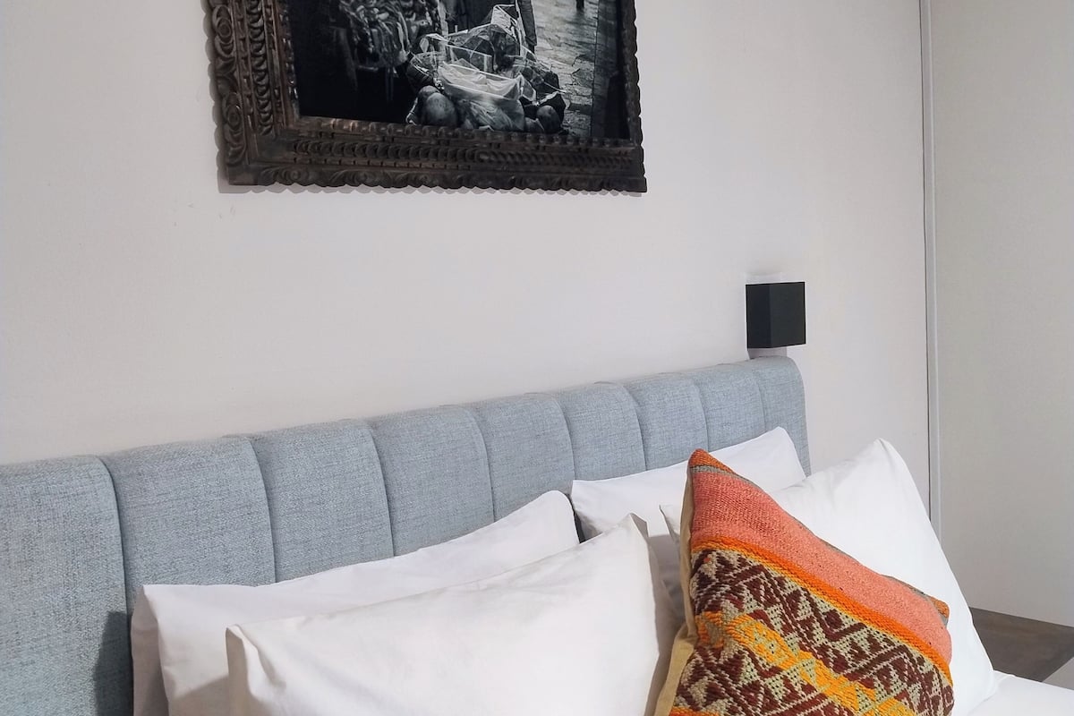 A bed with white and patterned pillows, a grey headboard, and a framed picture on the wall.