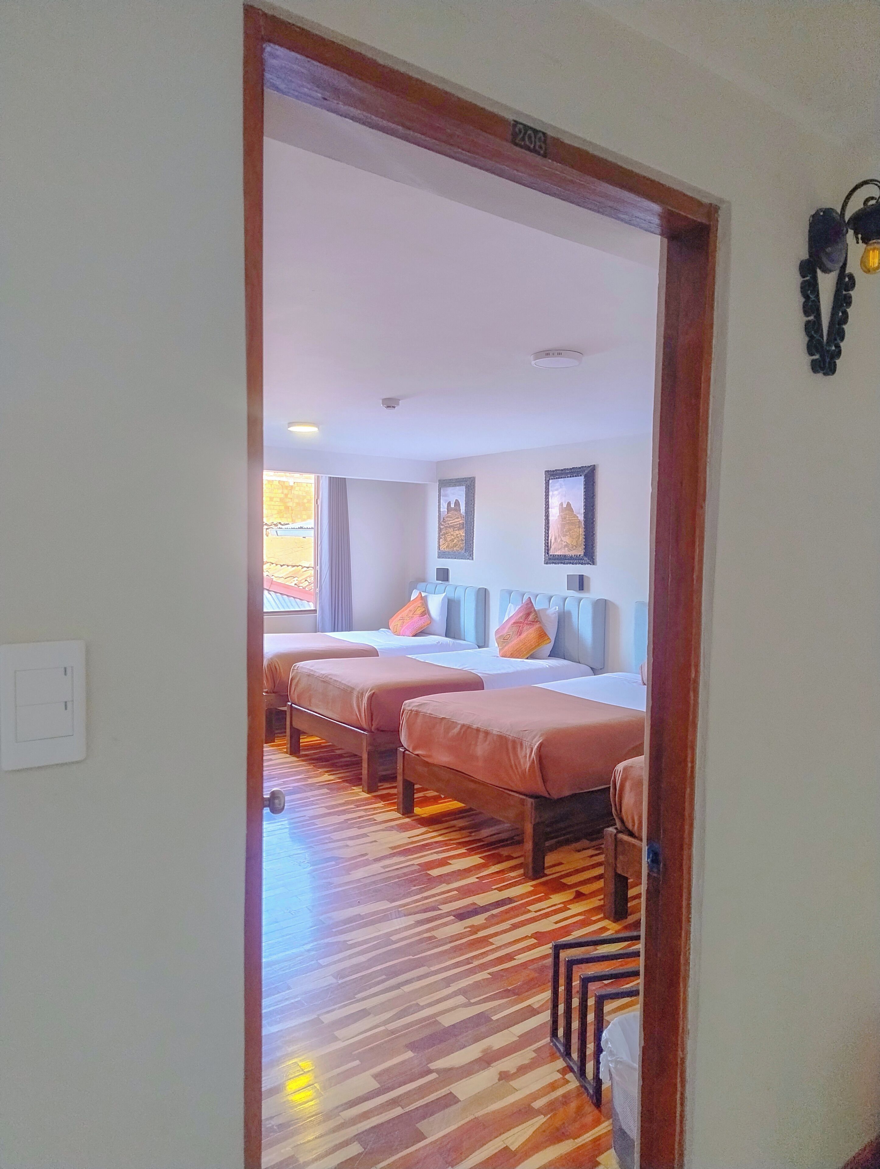 A hotel room with two beds, a wooden floor, a ceiling light, and a wall-mounted light fixture.