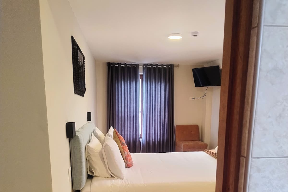 A hotel room with a wooden floor, a bed with white bedding, a television on the wall, and a window with curtains.