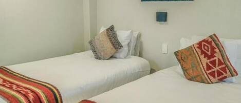 A hotel room with two beds, each with a white bedspread and a colorful blanket. There is a framed picture on the wall above the beds.