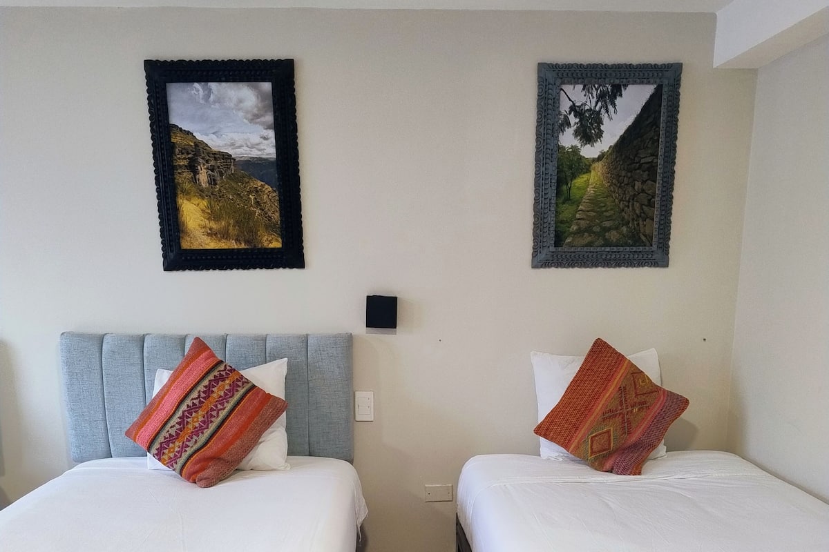 A room with two beds, each with a patterned pillow, a small wall-mounted light, and two framed pictures above the beds.