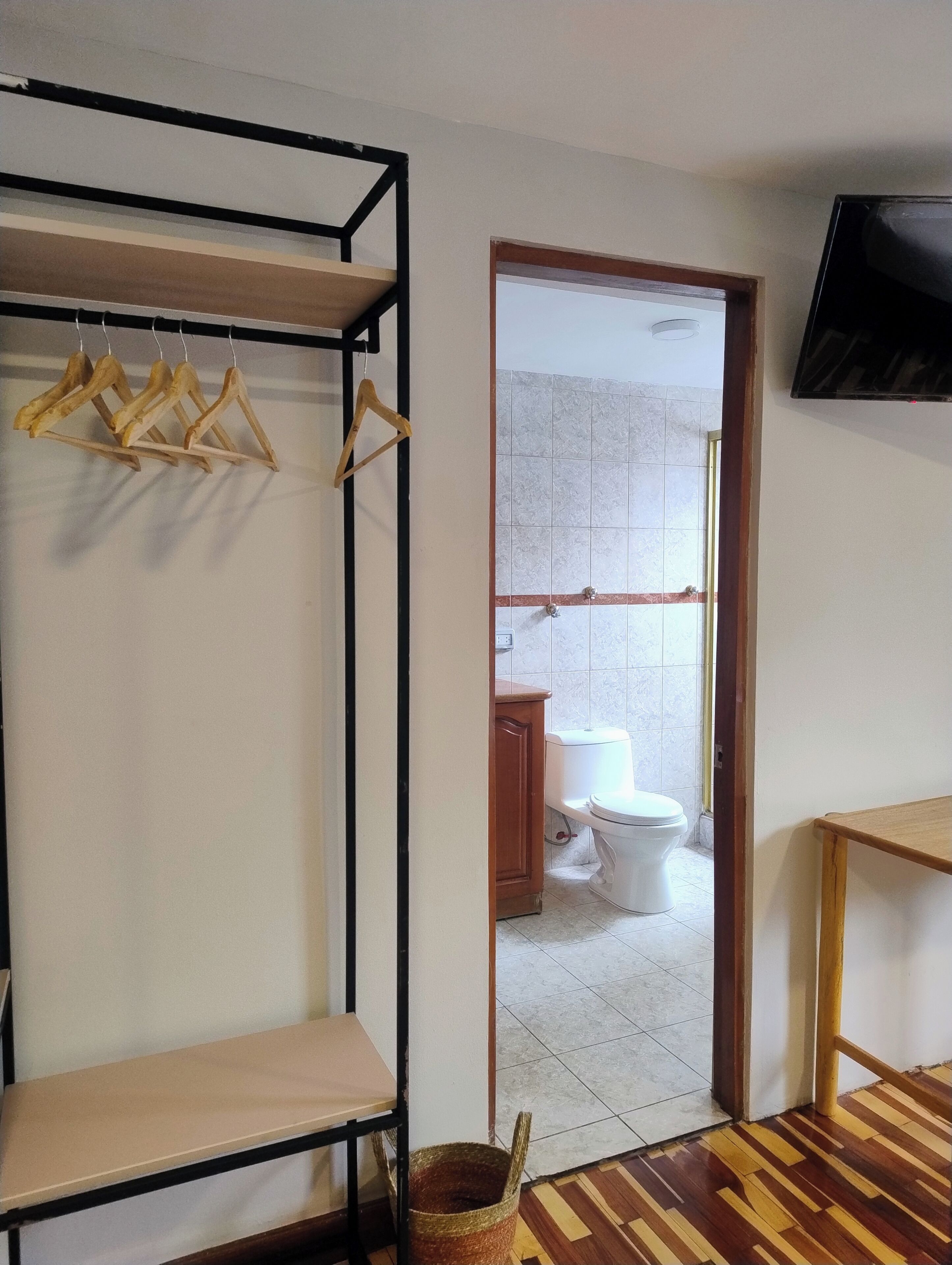 Una habitación con un armario de madera, un perchero, una mesa de madera y un televisor montado en la pared.