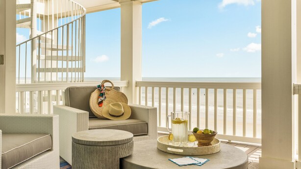 Un balcon agrémenté de meubles en osier, une table ronde avec une carafe et des fruits, et une vue sur la plage.