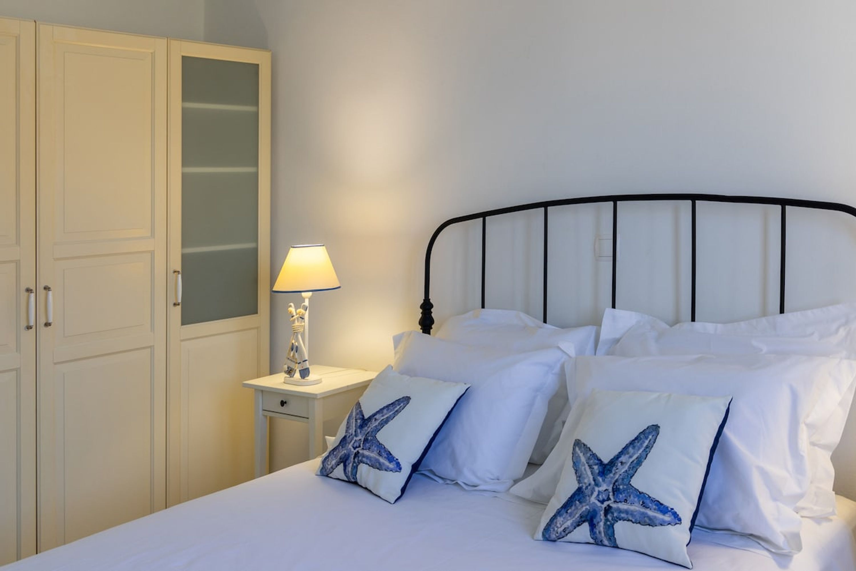 Una cama impecable con ropa de cama blanca y almohadas con forma de estrella de mar, una lámpara de noche y un armario blanco con puerta de cristal.