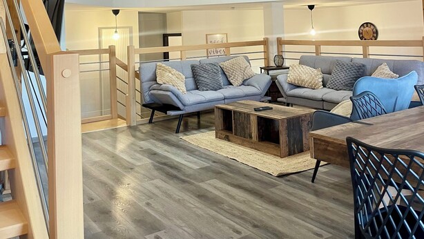Een ruime loft met een hoge plafond, zichtbare houten balken, een entresol met glazen hekwerk, een grijze zithoek en een houten eettafel.