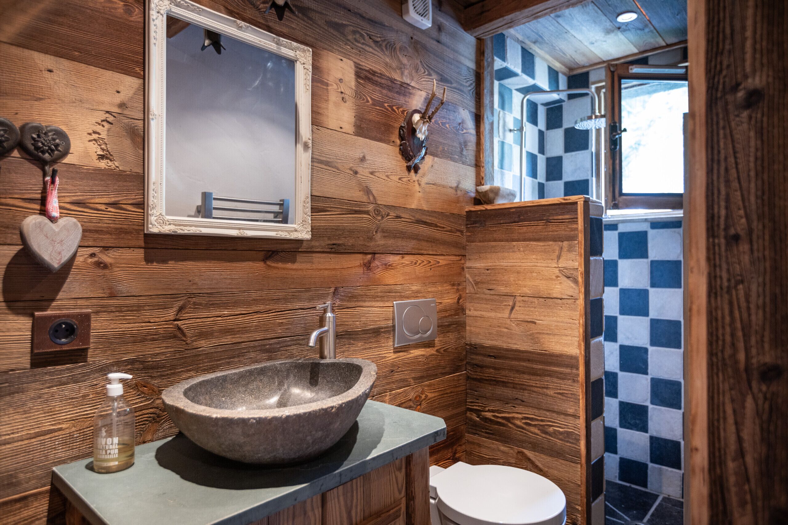 Un baño rústico con un lavabo de piedra, paredes de madera, un espejo y una zona de ducha a cuadros.