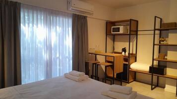 A hotel room with a bed, a desk, a chair, a microwave, and a shelf with a basket.