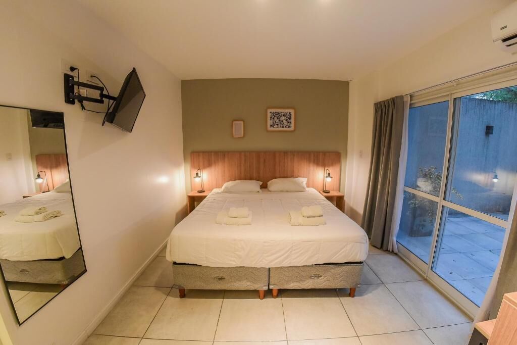 Habitación de hotel con una cama grande, un televisor de pantalla plana en la pared, una ventana con cortinas y una mesita con lámpara.