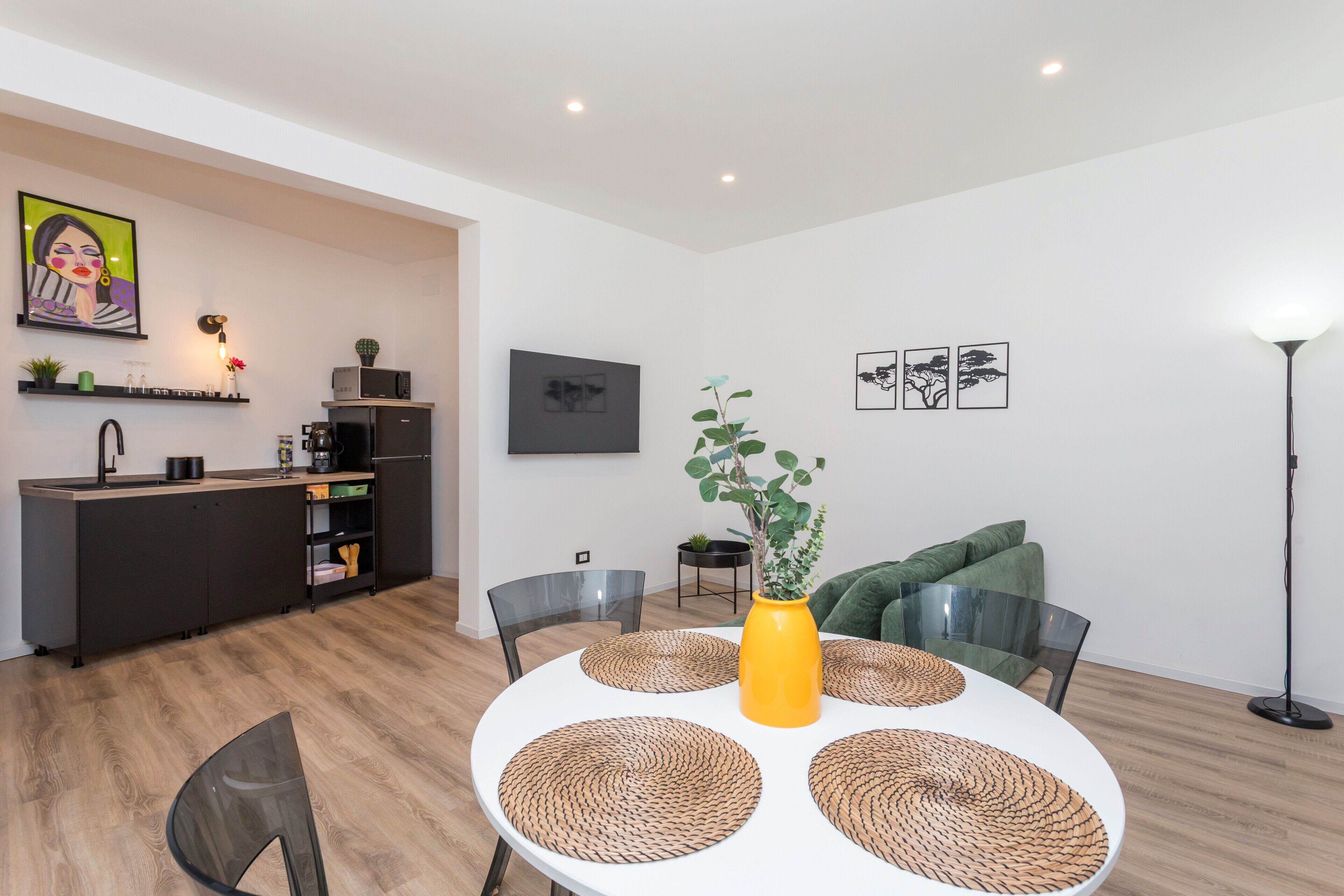 Een moderne keuken met een magnetron, koffieapparaat en een kleine potplant. Er is een eethoek met een witte tafel, gevlochten placemats en een gele vaas met een plant.