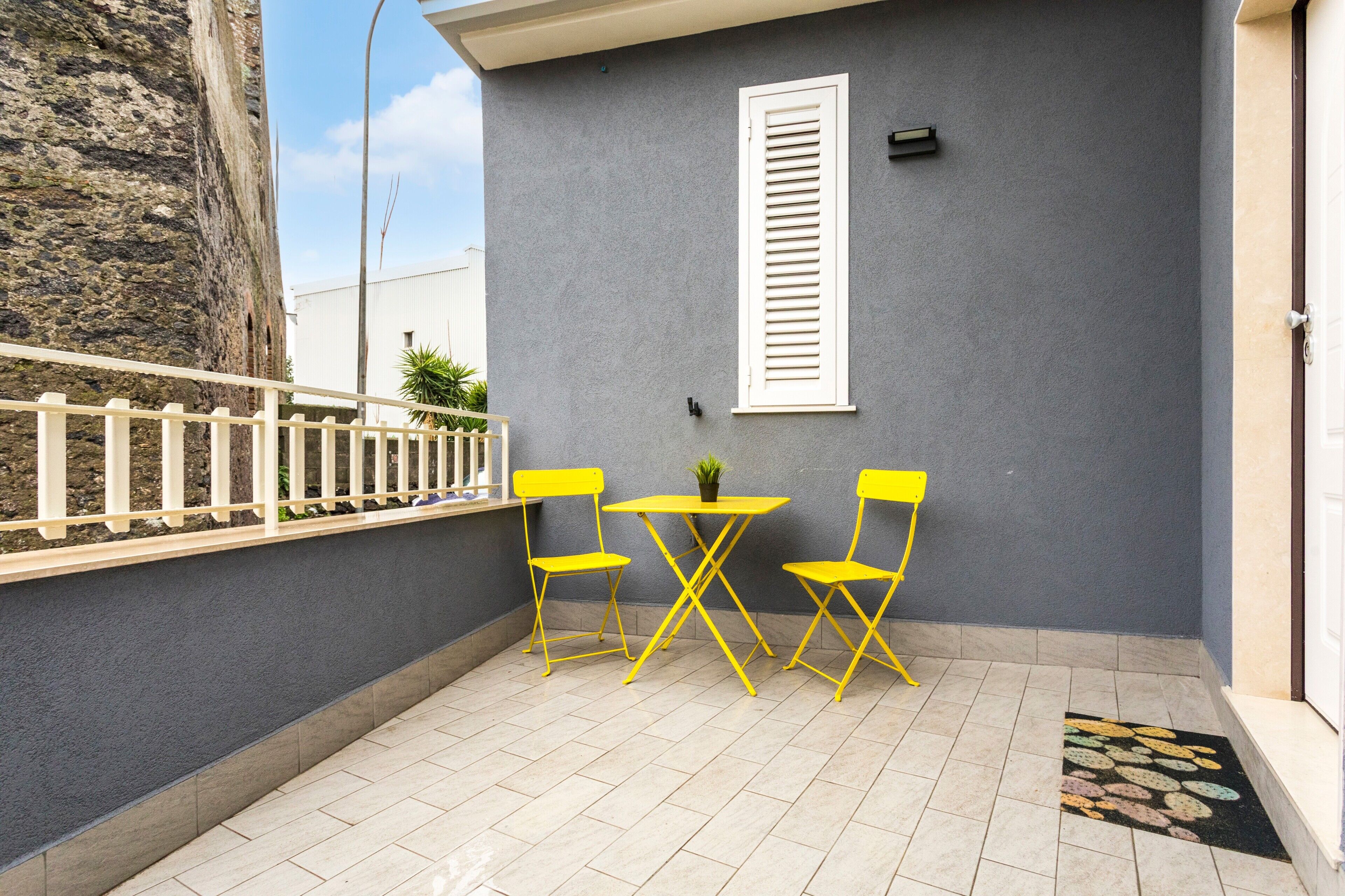 A balcony with a white fence, a grey wall, a white window shutter, and a tiled floor.