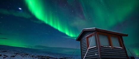 En træhytte med et vindue og en dør, beliggende på et snedækket område med en stjerneklar nattehimmel og nordlys i baggrunden.