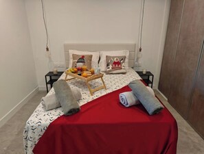 Ein ordentlich bezogenes Bett mit roter Tagesdecke, zwei Kissen und einer Obstschale.