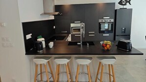 Eine moderne Küche mit dunklen Schränken, einer zentralen Kochinsel mit Hockern, einer Kaffeemaschine und einer Obstschale.