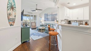 A spacious living area with a sofa, a coffee table, and a bar area with stools. There is a painting of a beach scene on the wall, and a ceiling fan is visible. - Coastal Canopy Modern Resort Condo w Massive Pool (North Myrtle Beach)