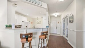A kitchen with white cabinets, a bar area with two stools, and a sign above the bar. - Coastal Canopy Modern Resort Condo w Massive Pool (North Myrtle Beach)