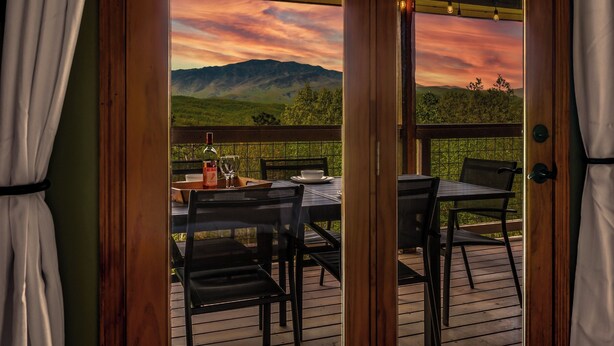 A wooden deck with a table set for two, overlooking a mountainous landscape with a sunset. - Private Location, Pet Friendly, Mountain Views, Game Area, Fire Pit + Hot Tub (Gatlinburg)