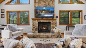 A living room with a stone fireplace, a television, and two large windows. - Private Location, Pet Friendly, Mountain Views, Game Area, Fire Pit + Hot Tub (Gatlinburg)