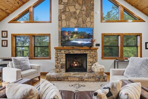 A living room with a stone fireplace, a television, and two large windows. - Private Location, Pet Friendly, Mountain Views, Game Area, Fire Pit + Hot Tub (Gatlinburg)