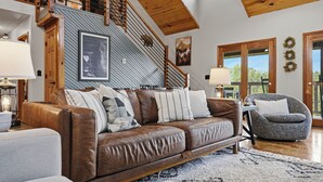 A spacious living area with a brown leather sofa, a grey armchair, and a staircase with wooden railings. - Private Location, Pet Friendly, Mountain Views, Game Area, Fire Pit + Hot Tub (Gatlinburg)