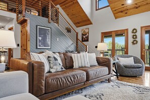 A spacious living area with a brown leather sofa, a grey armchair, and a staircase with wooden railings. - Private Location, Pet Friendly, Mountain Views, Game Area, Fire Pit + Hot Tub (Gatlinburg)