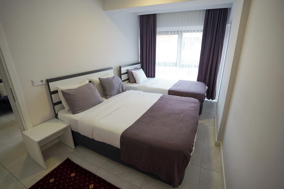 A hotel room with two beds, a nightstand, a small table, and a view of the outside through a window with curtains.