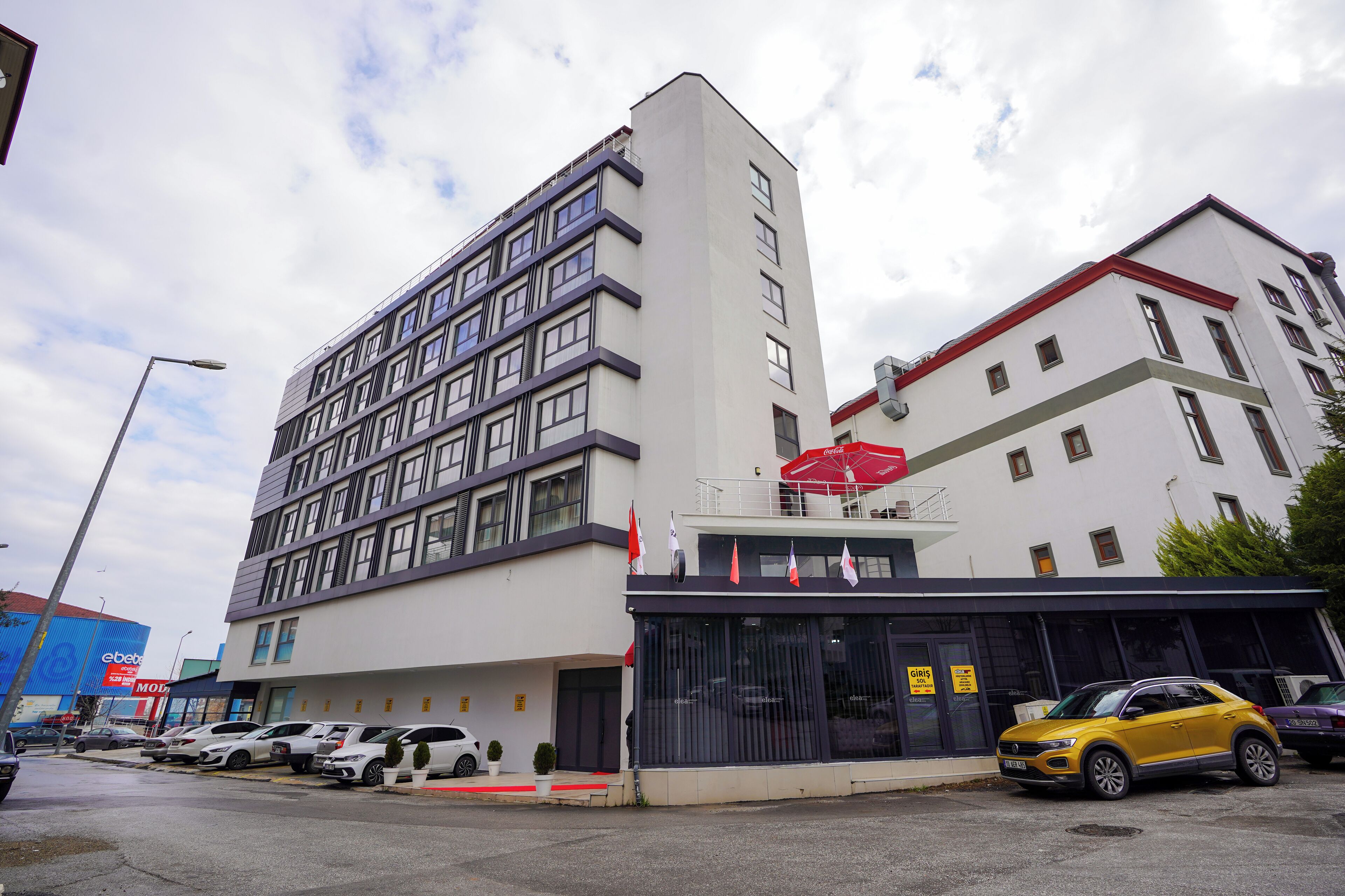 Un hotel de varios pisos con un diseño moderno, que cuenta con un estacionamiento con varios coches y una sombrilla roja cerca de la entrada.