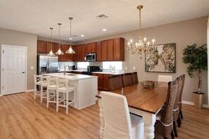A modern kitchen with wooden cabinets, stainless steel appliances, pendant lighting, and a dining area with a large wooden table and white chairs. - Spacious Home - Veranda Palms w/ Heated Pool & BBQ (Kissimmee)