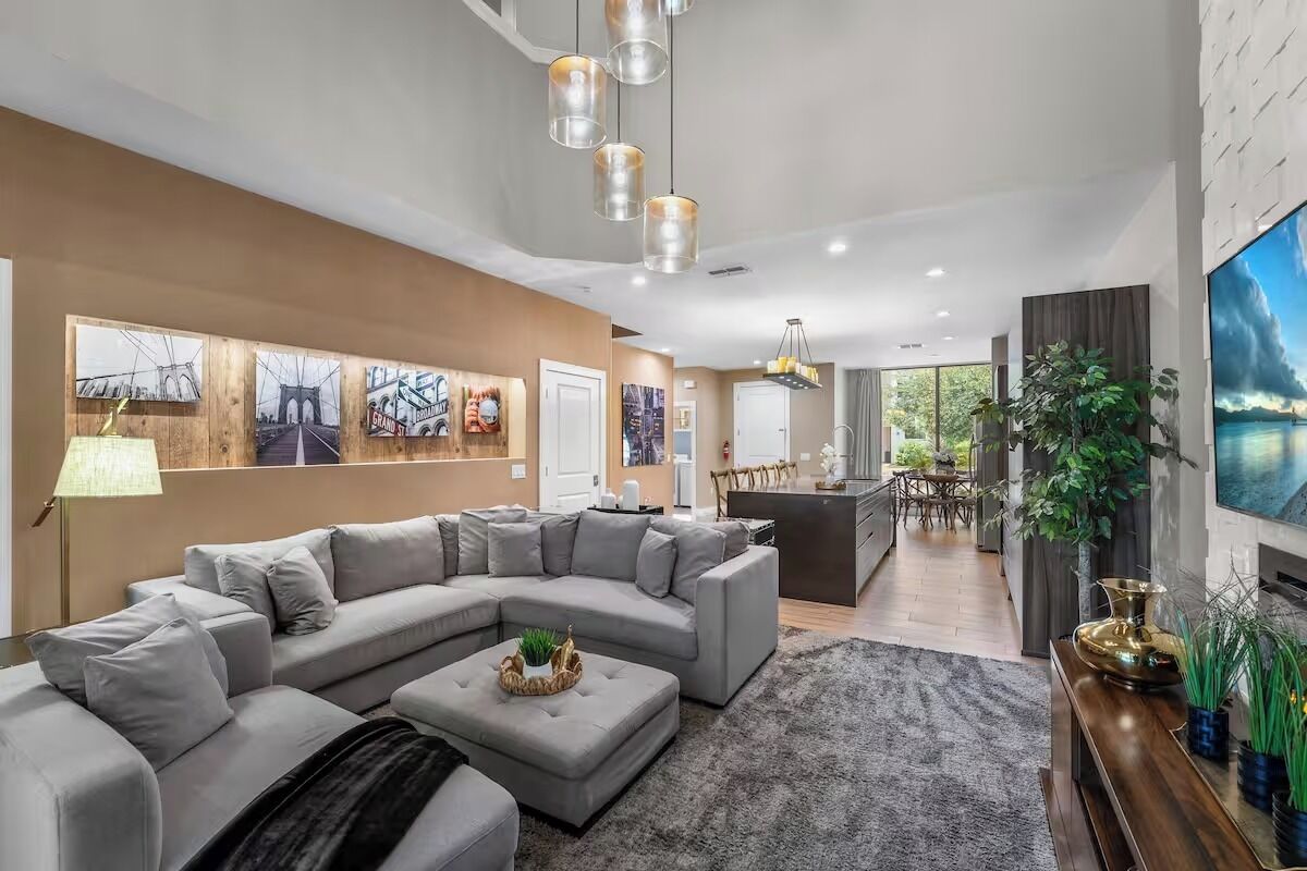 A modern living room with a sectional sofa, a coffee table, and a TV. There is a dining area with a table and chairs, and a kitchen in the background.