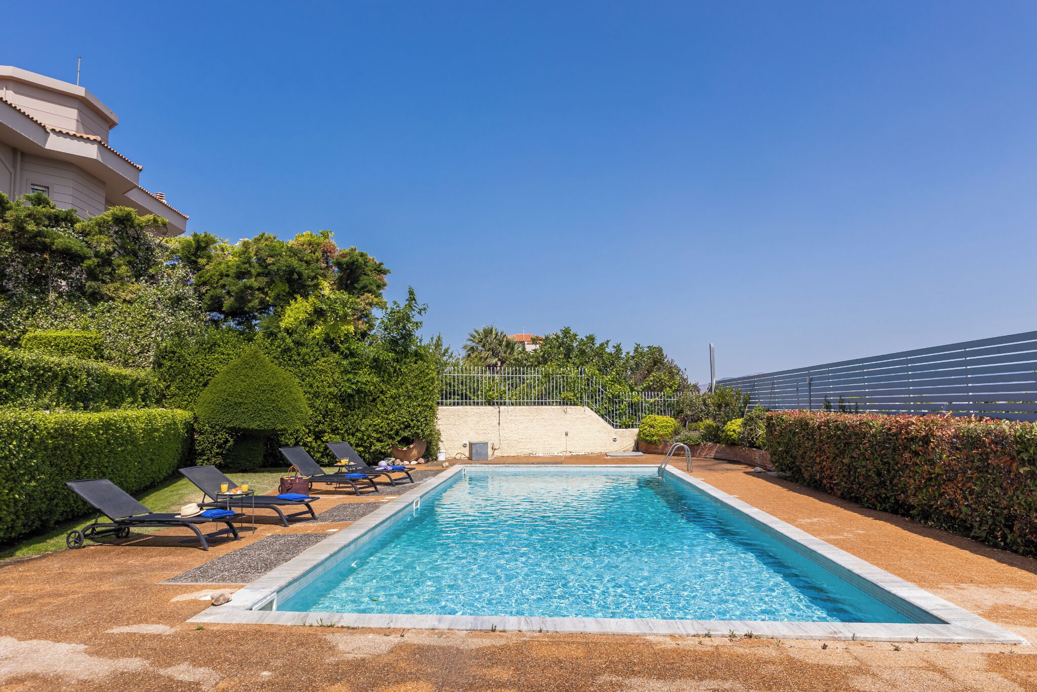 Une piscine rectangulaire à l’eau d’un bleu limpide, entourée d’une terrasse dallée. On y trouve des chaises longues avec des coussins bleus et une petite table sur laquelle sont posés une bouteille et un verre. L’espace piscine est bordé de haies et d’arbres soigneusement taillés.