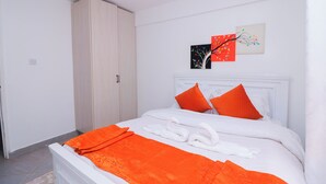Una cama bien tendida con ropa de cama naranja, almohadas blancas y un cabecero blanco. Se observan dos obras de arte en la pared sobre la cama, y un armario se ve a la izquierda.