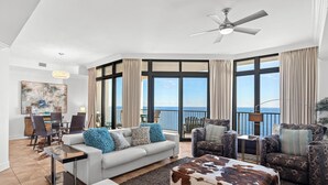 A spacious living room with a large window overlooking the sea, a ceiling fan, and a dining area with a view. - The Oasis At Orange Beach 2408 by Liquid Life (Orange Beach)