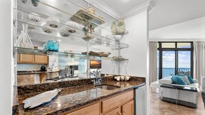 A modern kitchen with granite countertops, a stainless steel sink, and a glass display shelf above the counter. - The Oasis At Orange Beach 2408 by Liquid Life (Orange Beach)