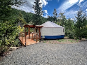 A yurt with a wooden deck and a covered porch, surrounded by a forested area.