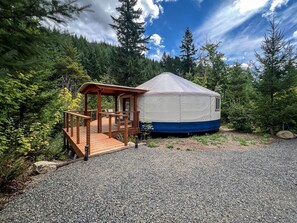 Une yourte dotée d’une terrasse en bois et d’un porche couvert, entourée d’une zone boisée.