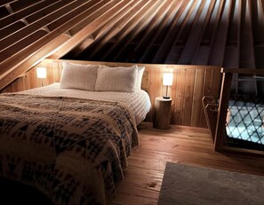 A bedroom with a sloped ceiling, a bed with a patterned blanket, two bedside lamps, and a window with a view. - Modern Forest Yurt with Firepit & Skylit Dome near Carson, Washington (Carson)