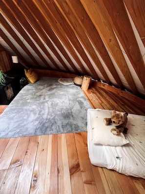 Dormitorio estilo loft con techo inclinado, vigas de madera, suelo que imita el mármol gris, colchón blanco, un osito de peluche y una planta pequeña.